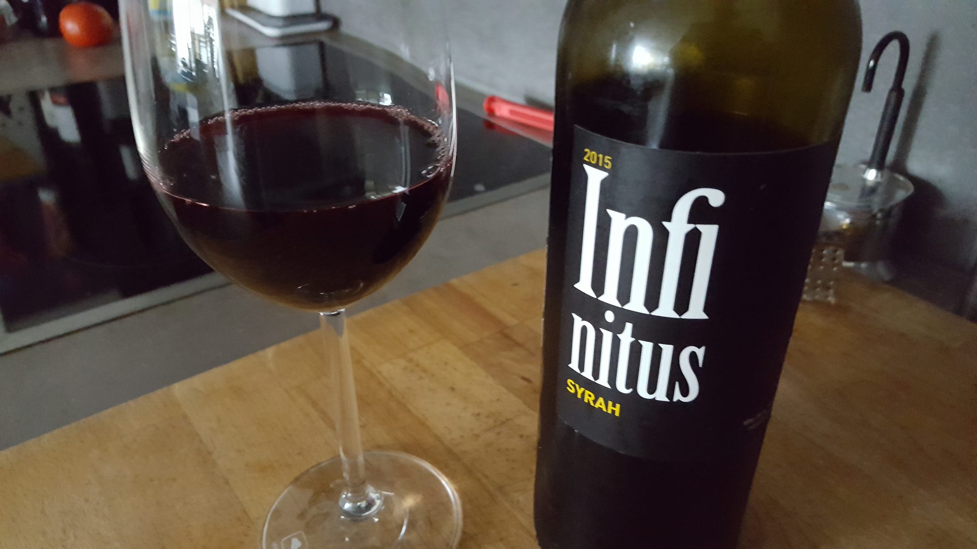 a bottle of 2015 infinitus syrah wine next to a filled glass on a wooden countertop the glass shows a deep red wine reflecting light while the bottle has a black label with white and yellow text digital production A bottle of 2015 Infinitus Syrah wine next to a filled glass on a wooden countertop. The glass shows a deep red wine, reflecting light, while the bottle has a black label with white and yellow text.