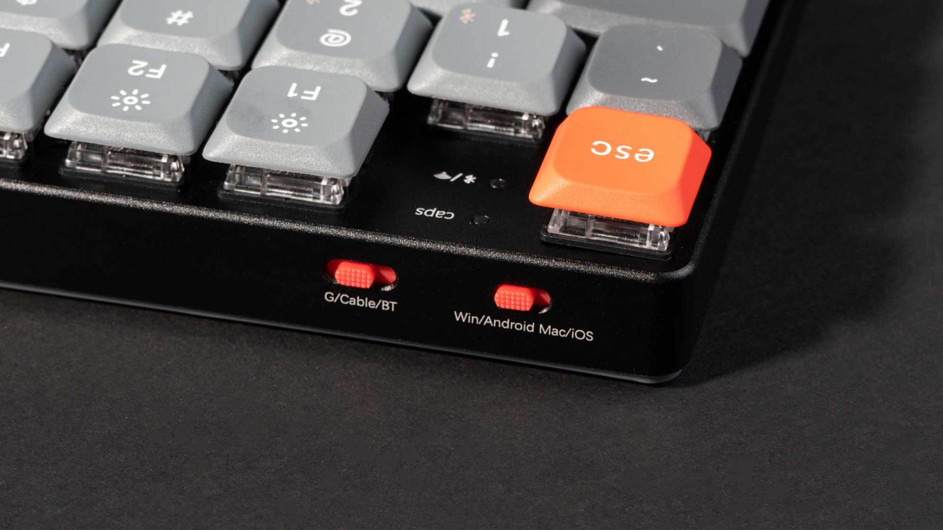closeup of a mechanical keyboard showing key switches and an orange esc key the side features toggle switches for cable mode and compatibility options labeled gcableit and winandroid macios digital production Close-up of a mechanical keyboard showing key switches and an orange ESC key. The side features toggle switches for cable mode and compatibility options labeled G/Cable/IT and Win/Android Mac/iOS.