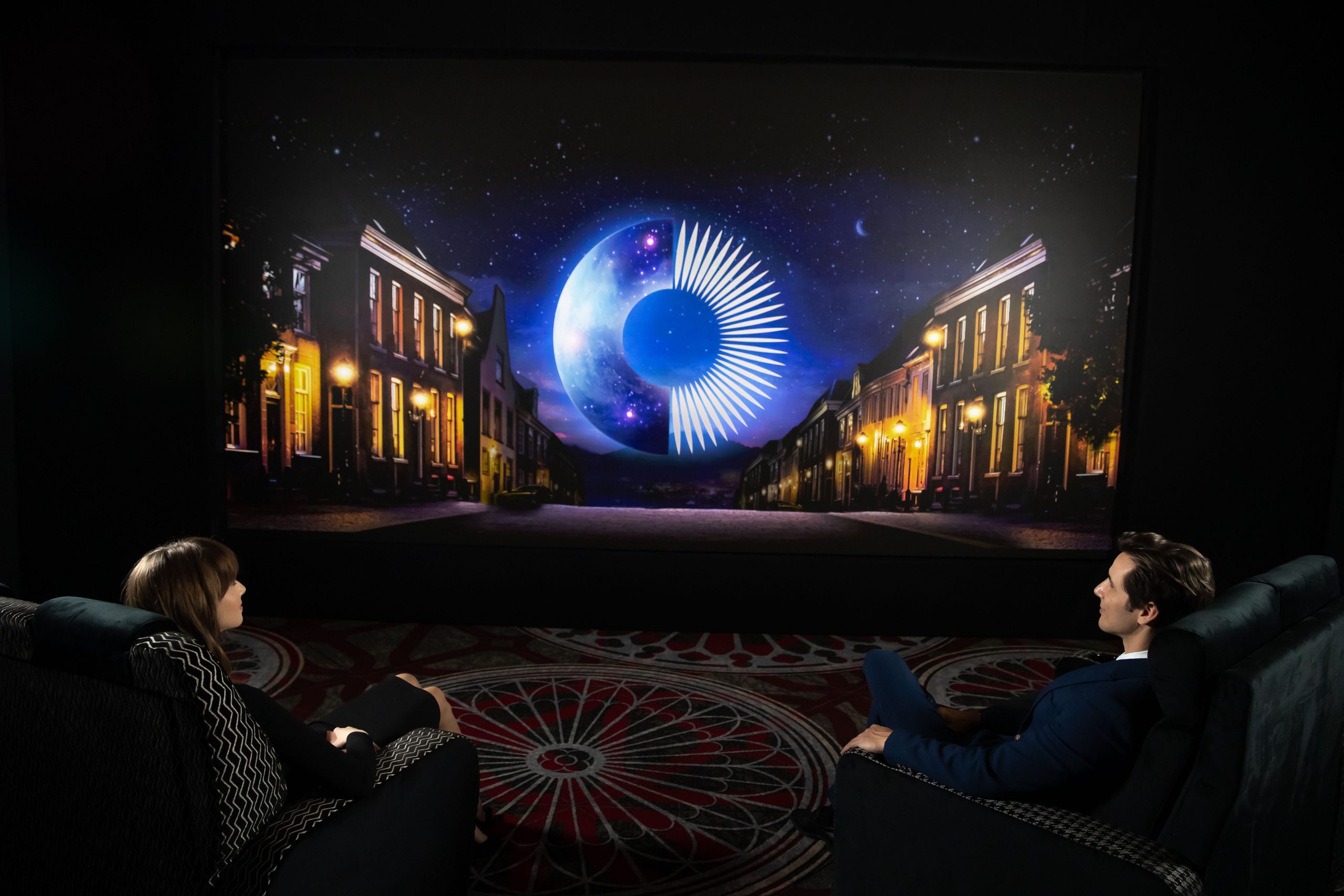 two people seated in plush chairs facing a large screen displaying a vibrant nighttime scene with a crescent moon and a stylized star explosion over a quiet street lined with illuminated buildings digital production Two people seated in plush chairs, facing a large screen displaying a vibrant nighttime scene with a crescent moon and a stylized star explosion over a quiet street lined with illuminated buildings.