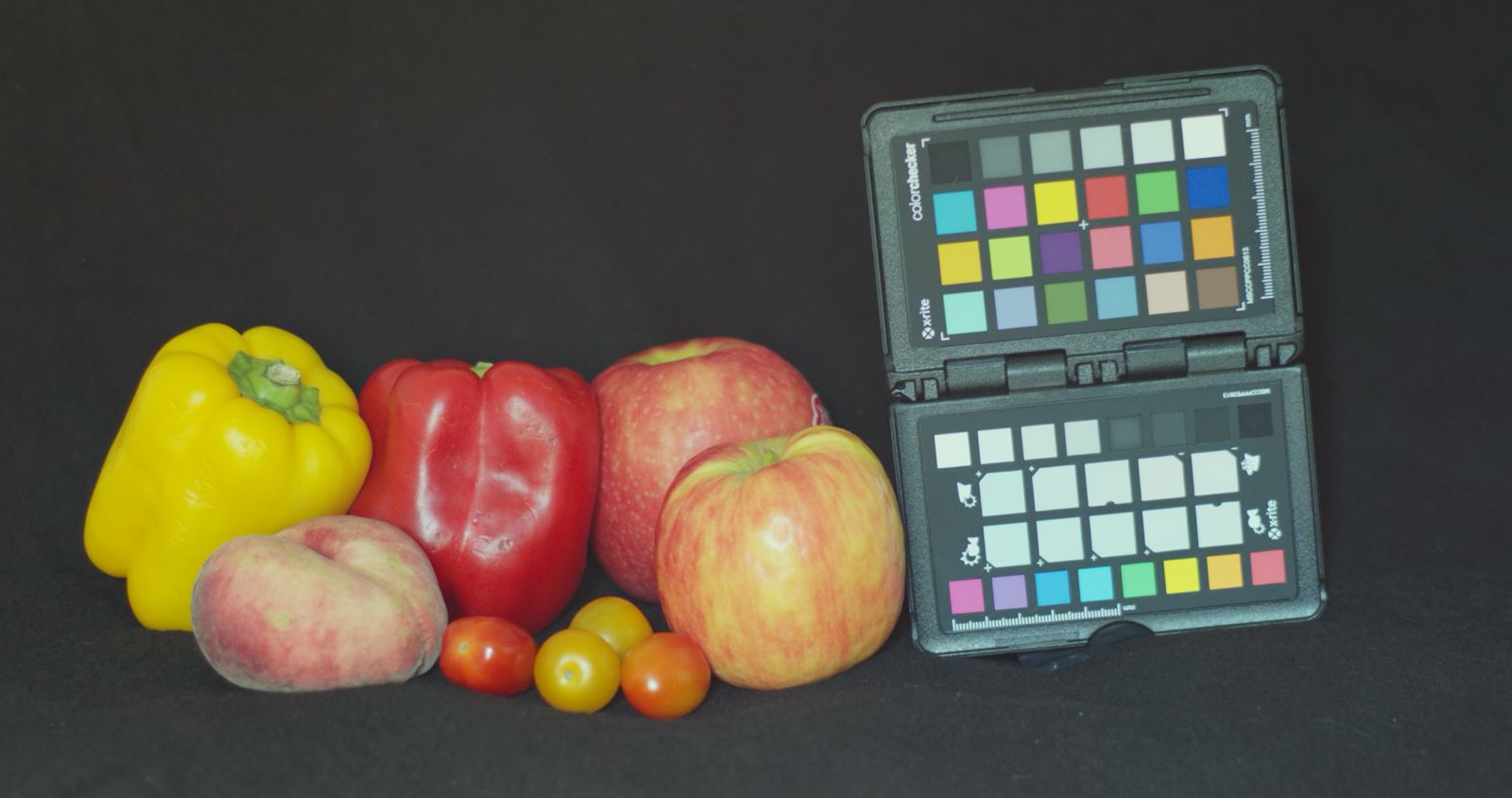 a collection of fresh fruits including a yellow bell pepper a red bell pepper two red apples a peach and several small cherry tomatoes arranged next to a color calibration chart on a dark background digital production A collection of fresh fruits including a yellow bell pepper, a red bell pepper, two red apples, a peach, and several small cherry tomatoes, arranged next to a color calibration chart on a dark background.